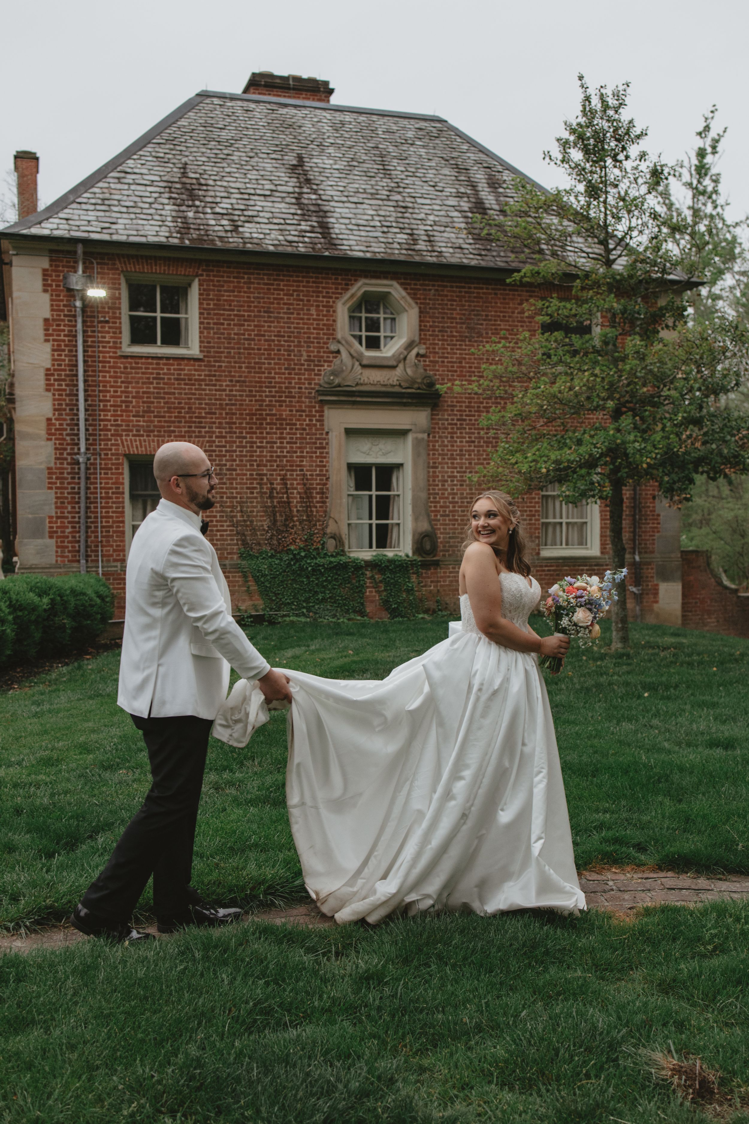 Bride and groom hold hands on green lawn before historic red brick mansion on their wedding day.