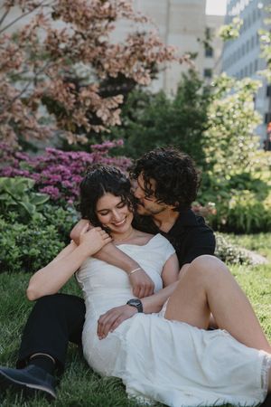 A bride in a white dress smiles as her groom kisses her cheek while sitting on grass in a lush garden.