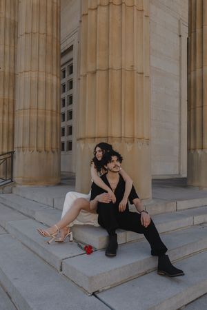 Couple sitting on stone steps of a neoclassical building, woman in white dress embracing man in black suit with a red rose nearby.