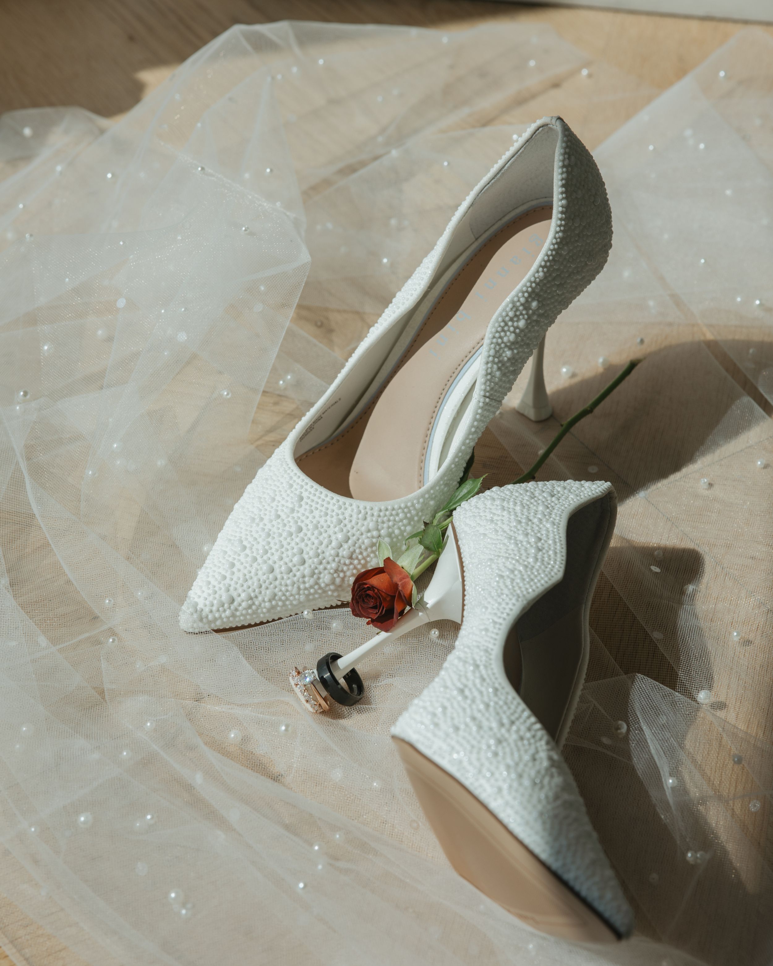 White lace bridal heels with a delicate red rose and wedding rings displayed on sheer tulle fabric.