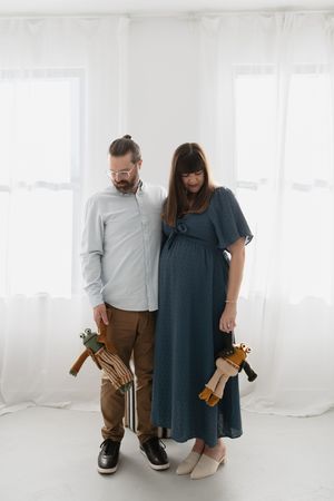 A couple stands together holding wooden hangers in a bright white studio setting, wearing casual yet stylish outfits.