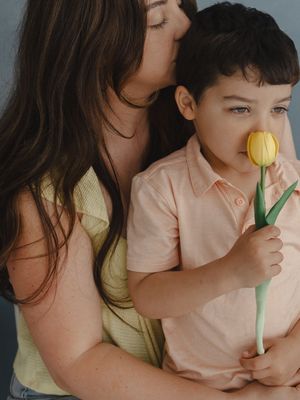 A mother kisses her young son as he smells a yellow tulip flower, both wearing pastel clothing.