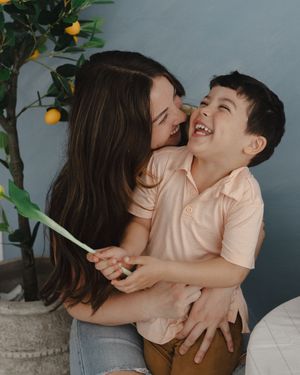 A woman with long dark hair hugs and laughs with a young boy in a pink polo shirt near a lemon tree.