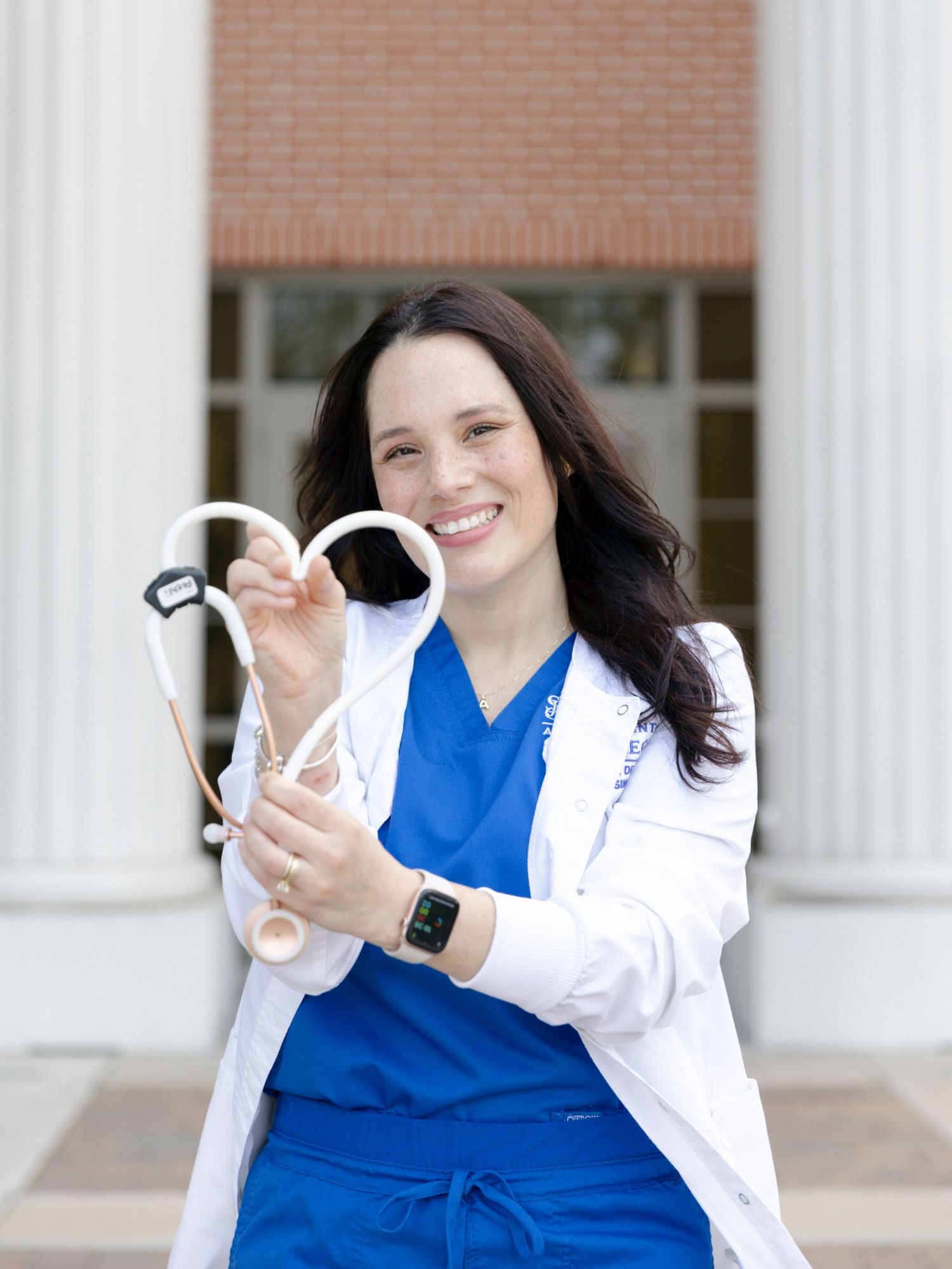 Medical professional in blue scrubs and white coat smiles while standing in front of a building with columns.