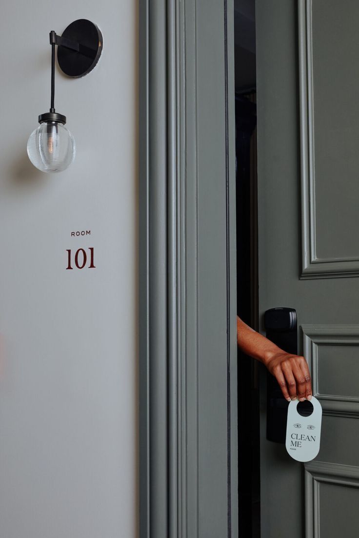 A hand reaches through a hotel door to hang a 'Clean' sign on room 101, with a modern spherical wall sconce nearby.