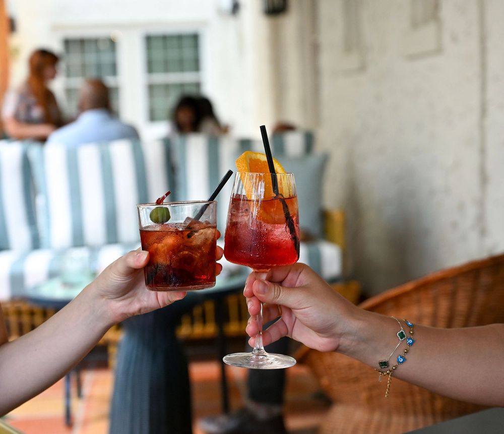 Two hands toast with colorful cocktails garnished with cherries at a restaurant patio setting.