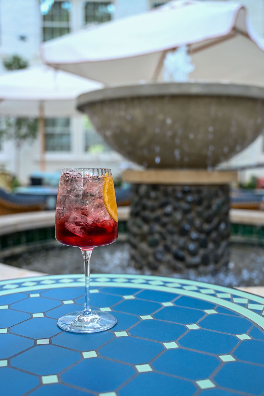 A red cocktail in a wine glass sits on a blue mosaic table with a fountain in the background.