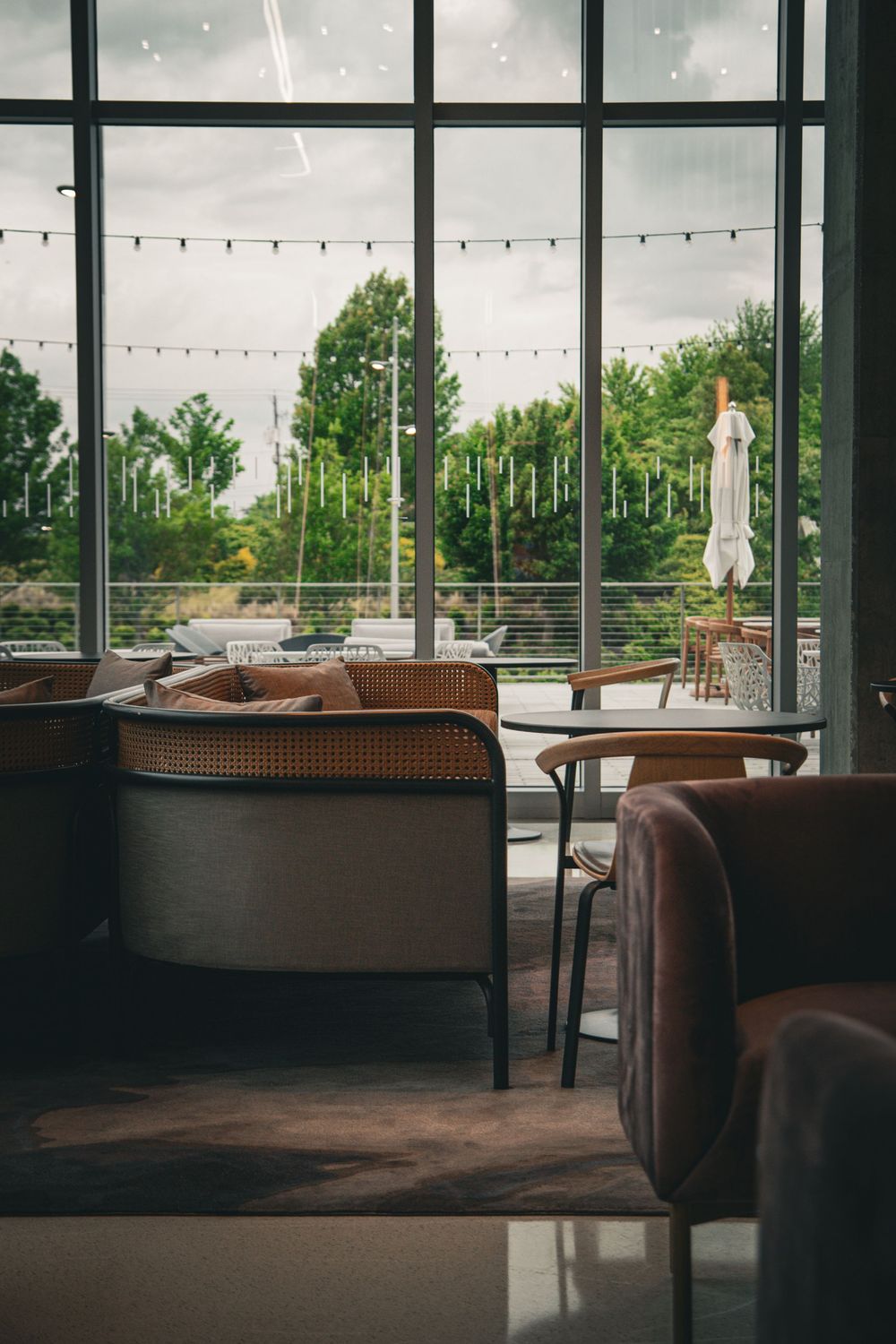 Modern restaurant interior with panoramic windows, string lights, and comfortable brown seating overlooking an outdoor area.