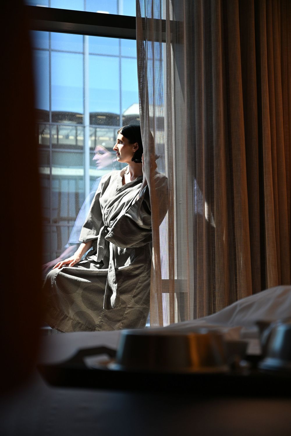 Woman in gray robe standing by sheer curtains gazing out a sunlit hotel window with city views.