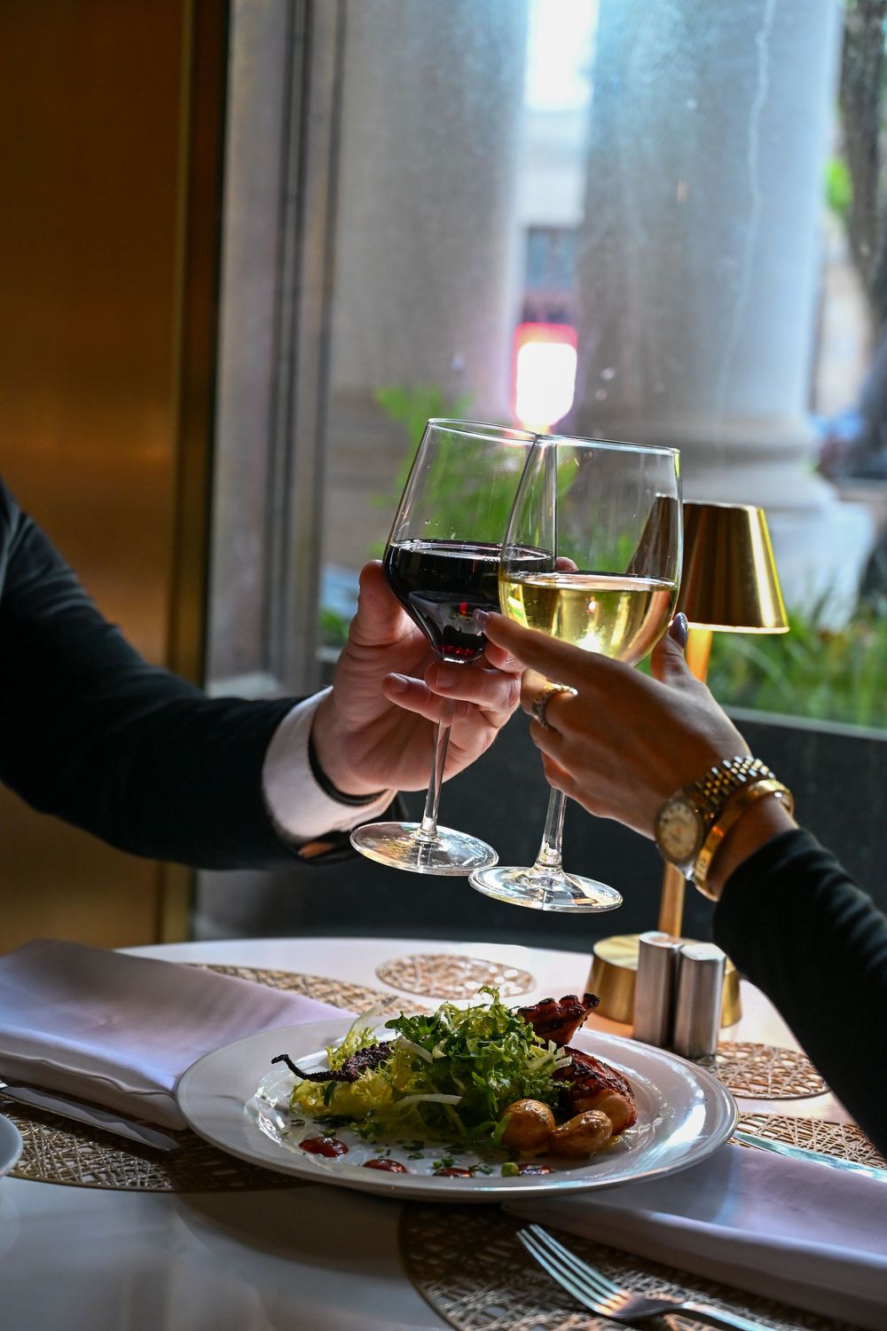Two people toast with glasses of red and white wine over an elegantly plated meal at a restaurant table.
