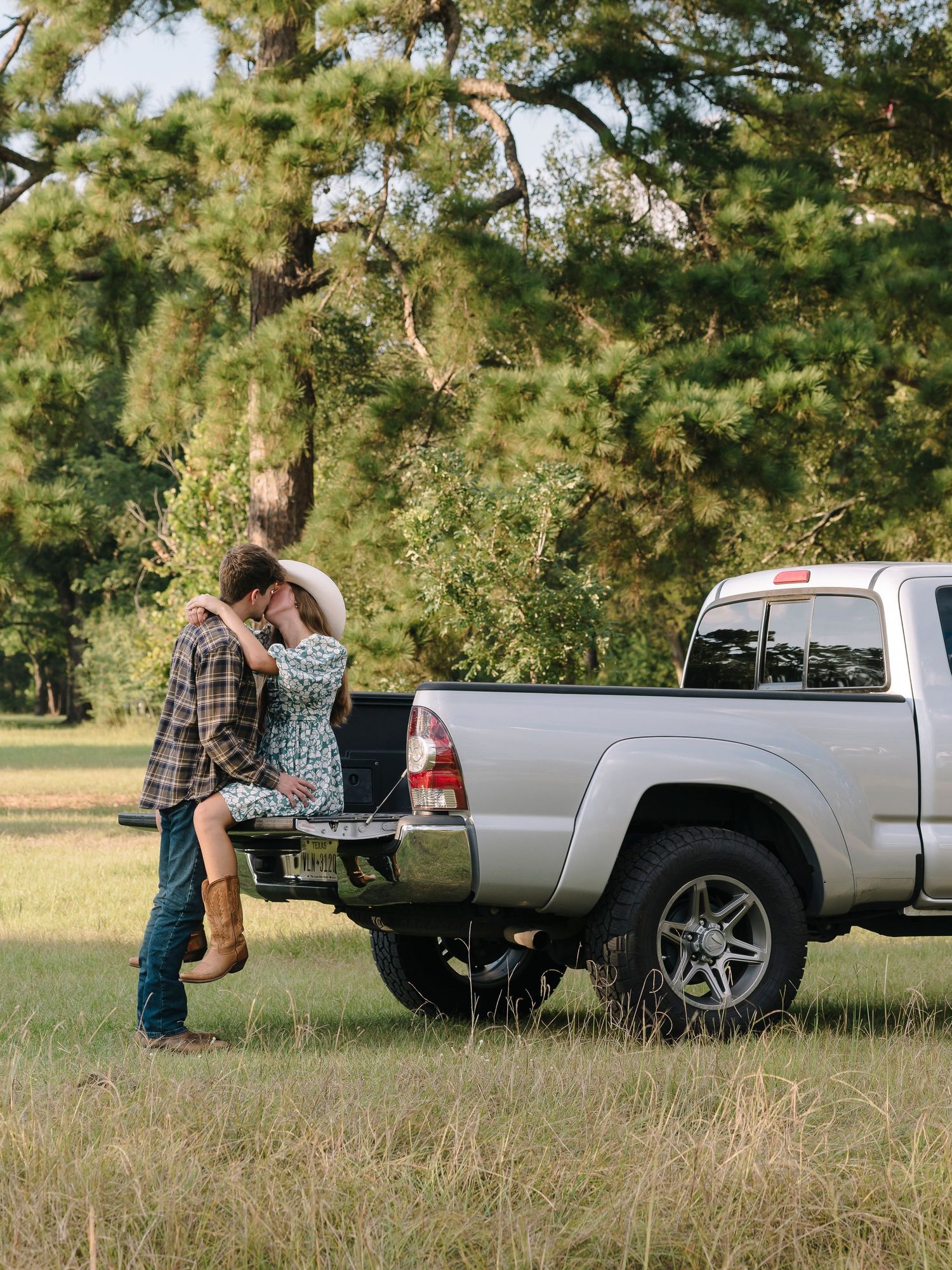A couple shares a romantic moment by a silver pickup truck tailgate in a scenic grassy field with pine trees in the background.