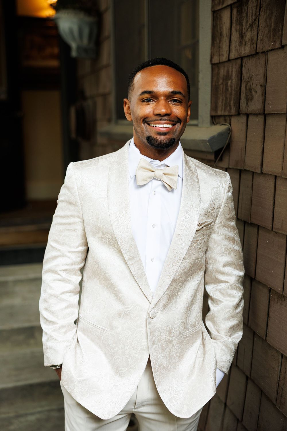 A groom in a pristine white tuxedo with bow tie smiles warmly while standing against a rustic stone wall.