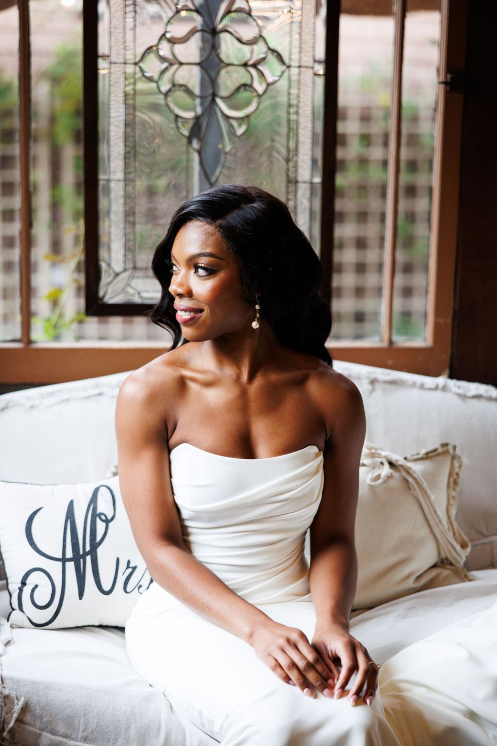 Strapless white gown adorns bride on cream colored couch near decorative window at Chapel of Orange before she walks down the aisle.