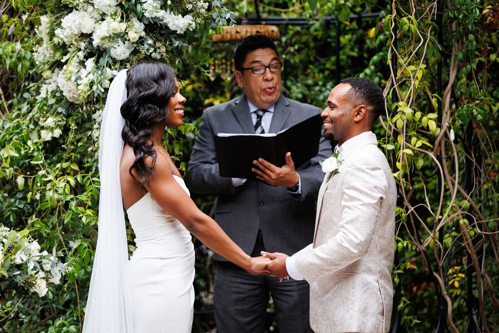 Wedding ceremony taking place outdoors with couple holding hands at the altar in front of officiant under floral arch.