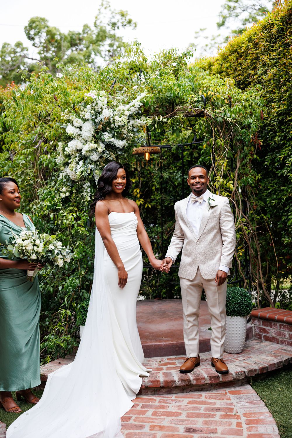 A wedding couple stands together on a garden patio with lush greenery and white floral arrangements in the background.
