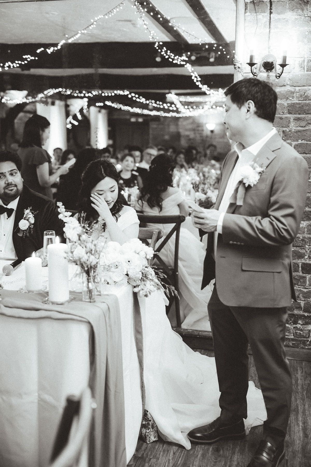 Black and white photo showing guests at a reception socializing near cocktail tables decorated with white flowers and twinkling lights.