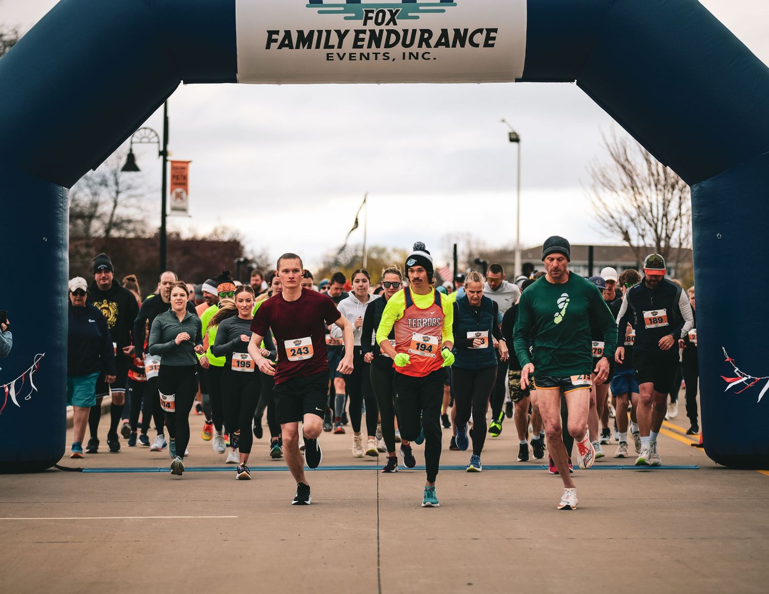 Runners line up at the starting line under a black inflatable arch at an outdoor endurance race event.