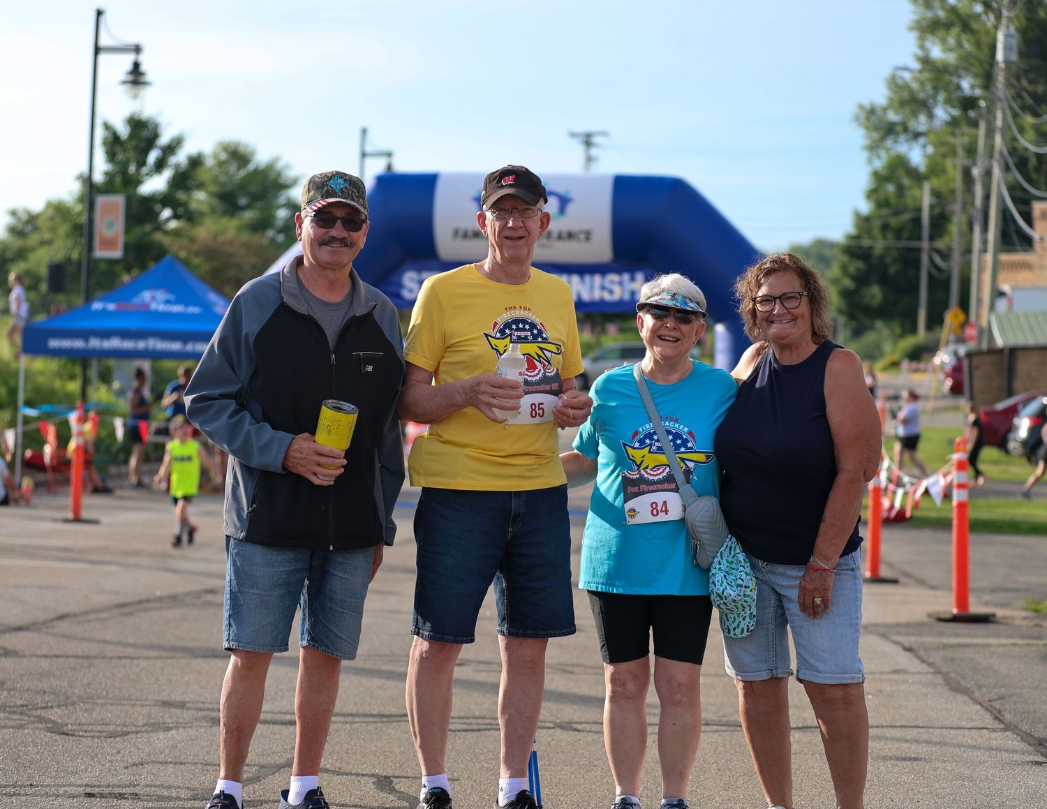 A group of runners pose together at a summer race event with orange cones and a blue inflatable arch in the background.