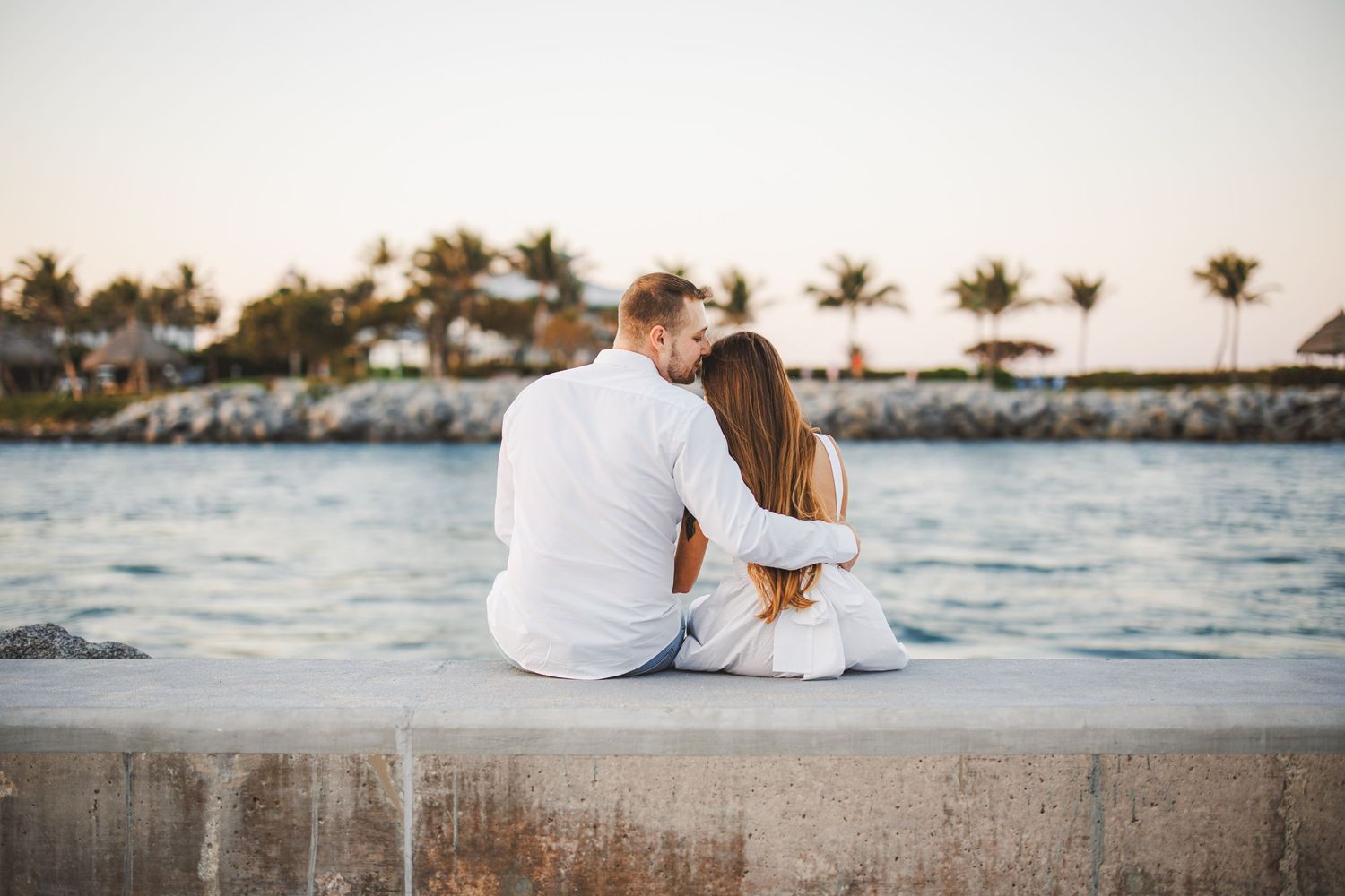 Couple in white outfits sitting together on rocky beach at sunset in romantic tropical setting.