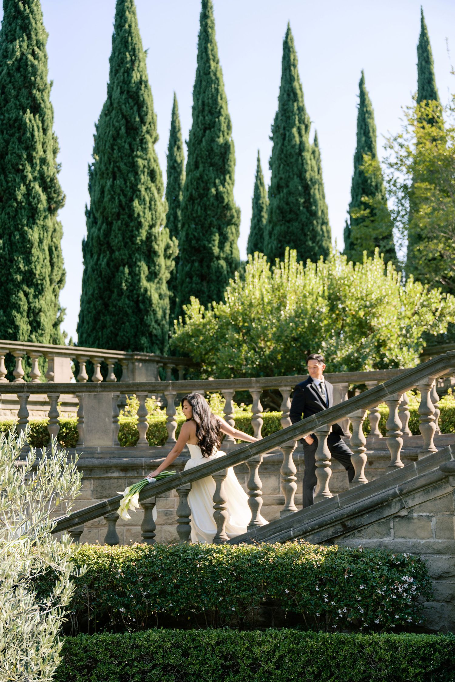 Stone balustrade with ornate balusters lined by tall cypress trees in a Mediterranean garden setting.