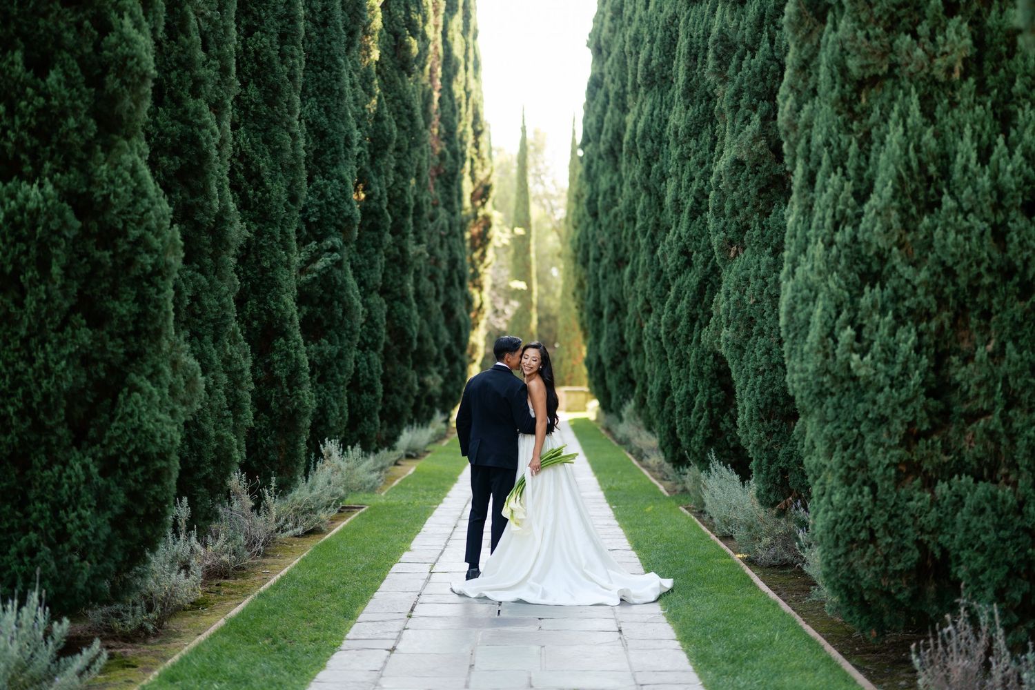 Couple walks down a romantic cypress tree-lined path in elegant wedding attire during outdoor ceremony.