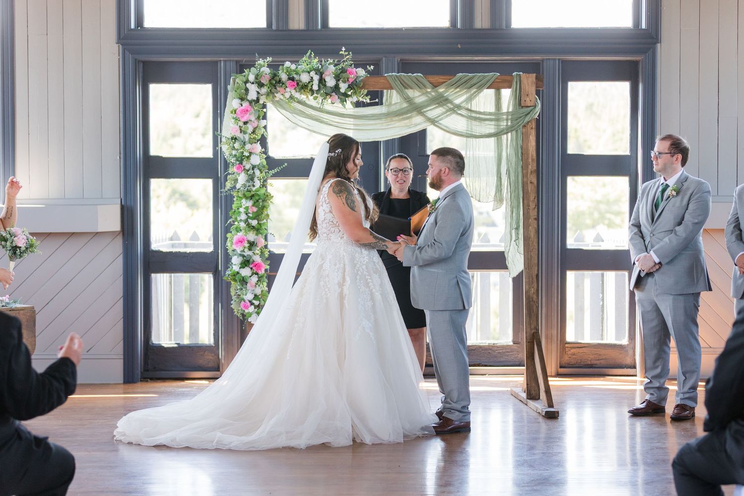 A wedding ceremony takes place under a floral arch in a bright sunlit venue with wooden floors and glass doors.