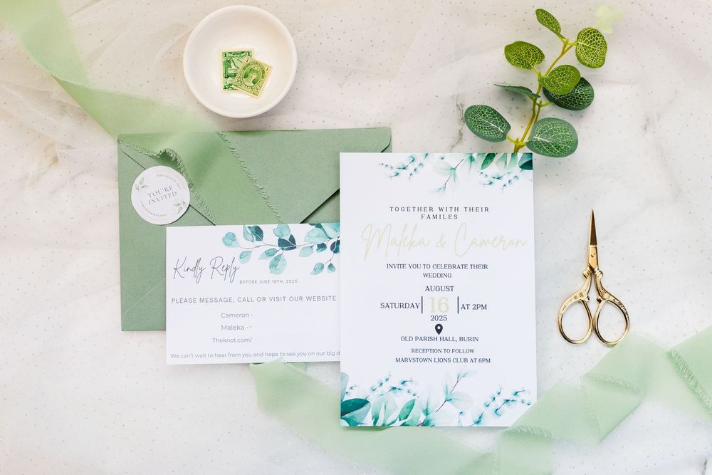 Wedding invitation suite in white and sage green with watercolor eucalyptus design, ribbon and gold scissors on marble surface.