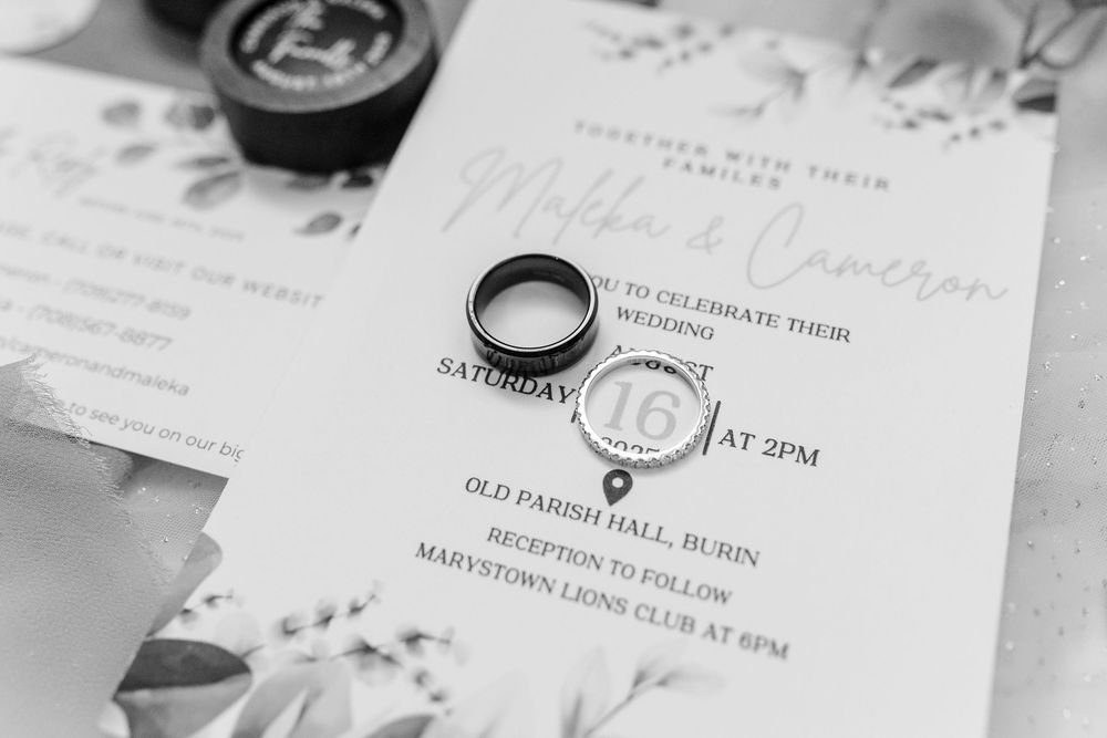 A black and white photo of wedding rings placed on a wedding invitation with elegant lettering and floral details.