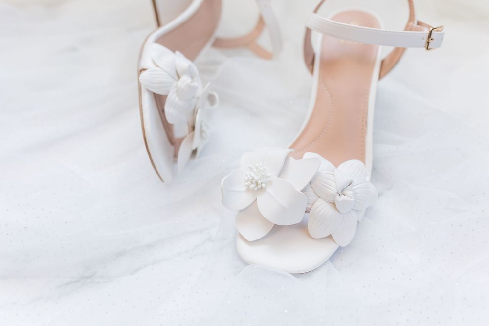 Elegant white bridal sandals with delicate floral appliques and ankle straps on a light background.