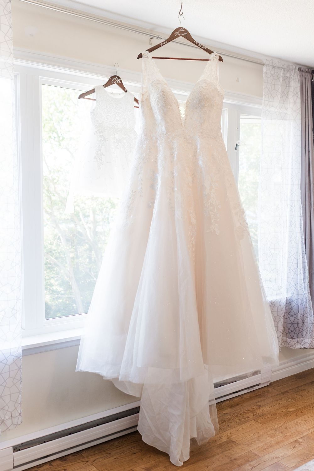 Two elegant white wedding dresses hanging by wooden hangers against a sunlit window in a bright room.