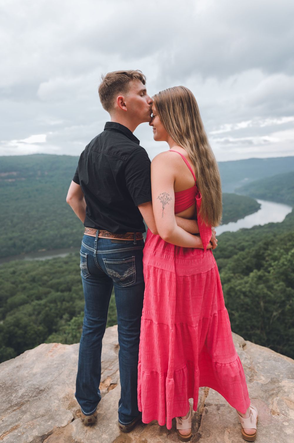 Couple sharing an intimate moment on a rocky overlook with winding river and green valley views below cloudy skies.