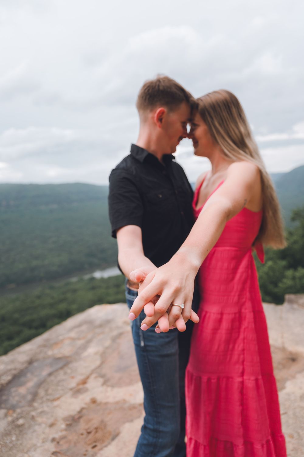 A couple shares an intimate moment on a mountain overlook, with the woman wearing a flowing red dress.