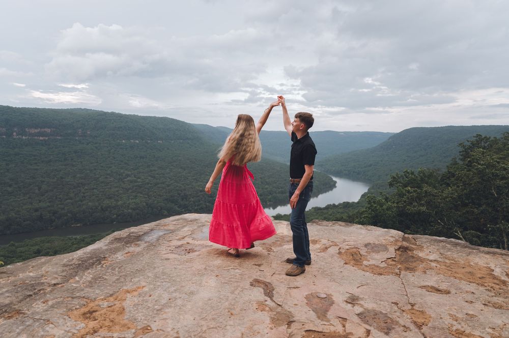 Two people standing on a rocky mountaintop overlook with sweeping valley and river views below under cloudy skies.