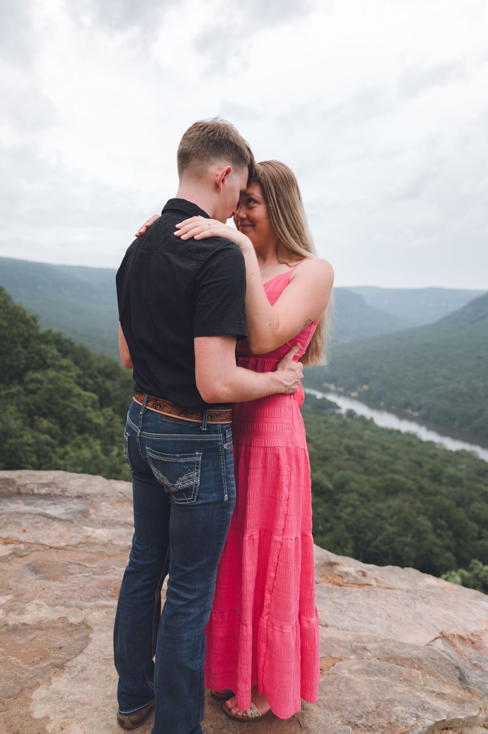A couple embraces on a mountain overlook with scenic views of rolling hills and valleys in the background.