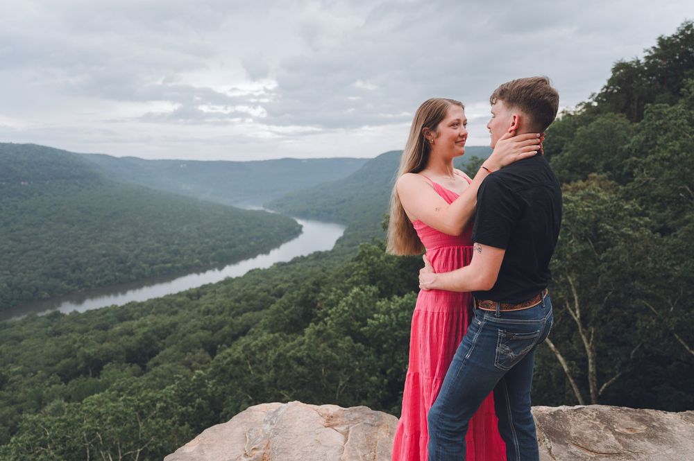 Romantic couple in formal attire embraces while enjoying scenic mountain overlook at a river valley on a cloudy day.