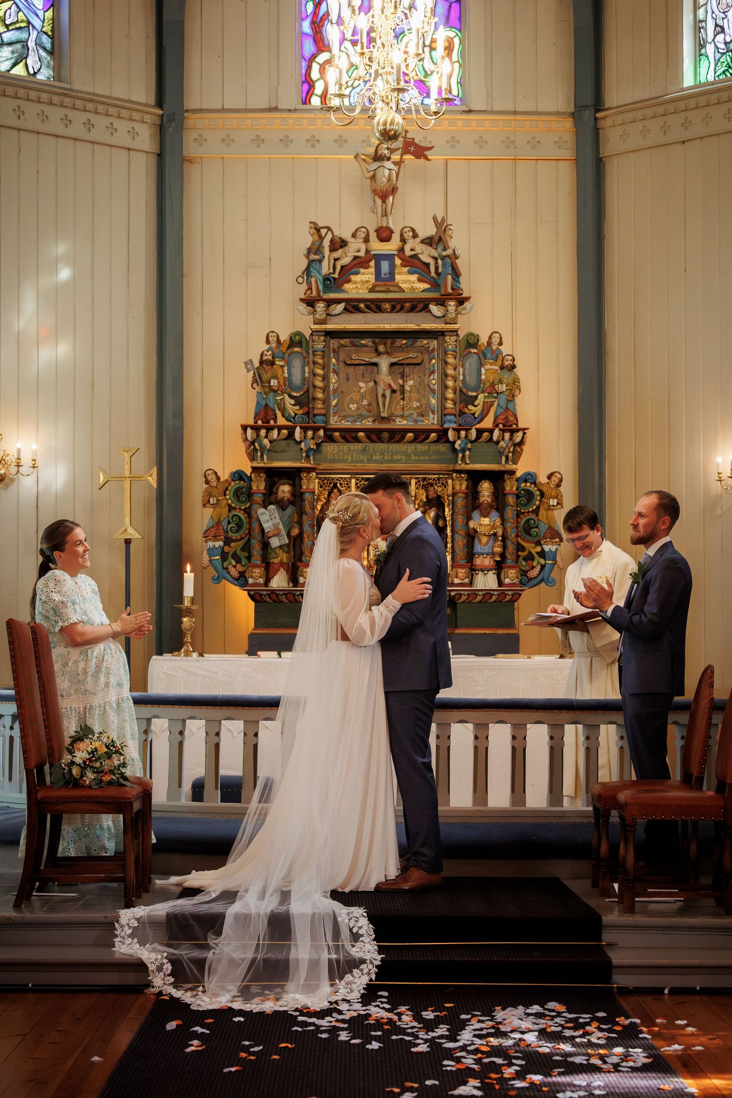 A wedding ceremony takes place in front of an ornate golden altar in a historic church with stained glass windows and flower petals.