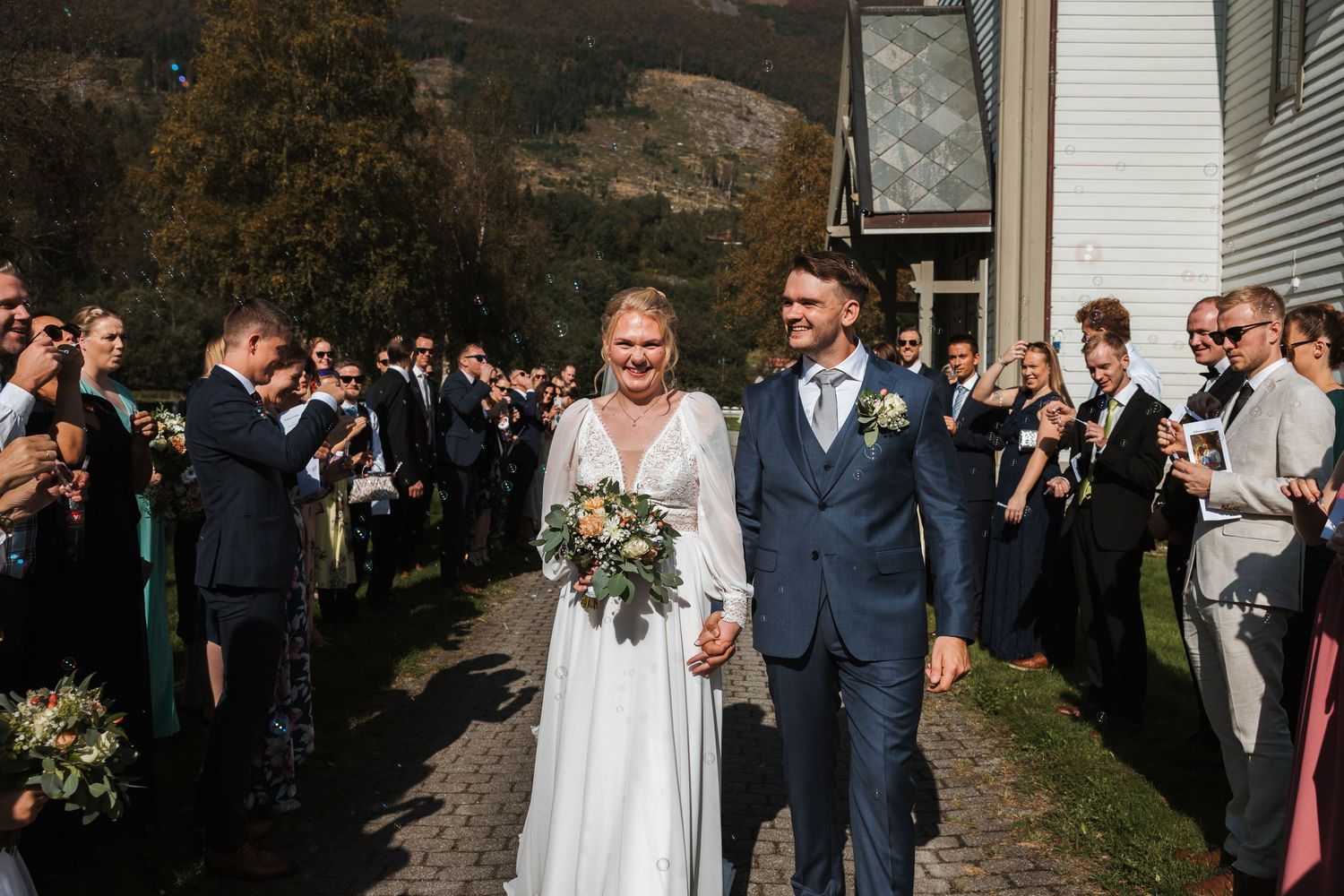 Wedding party and guests walk along pathway outside white building during outdoor celebration.