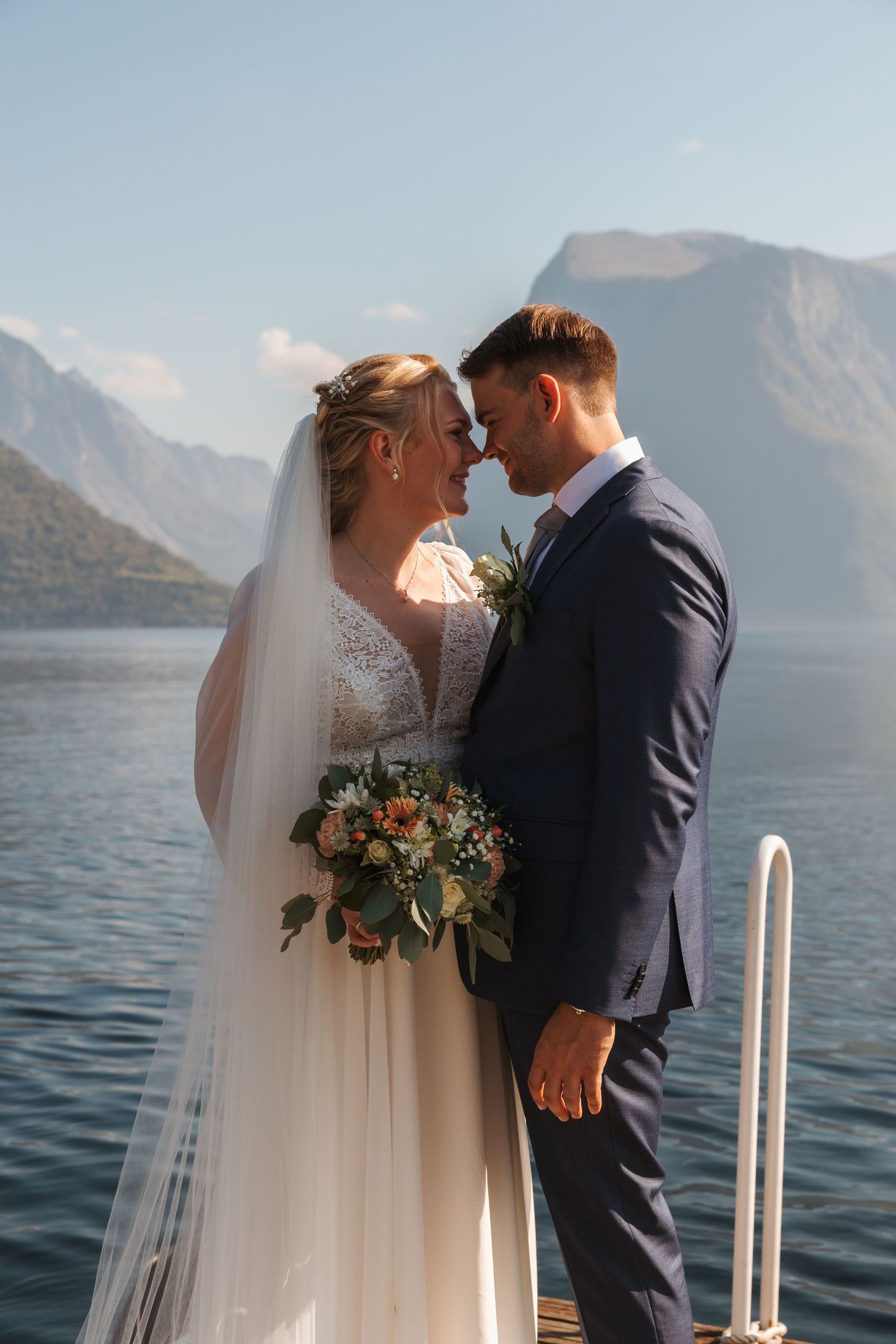 A romantic wedding couple embraces on a dock with stunning mountain lake scenery in the background.