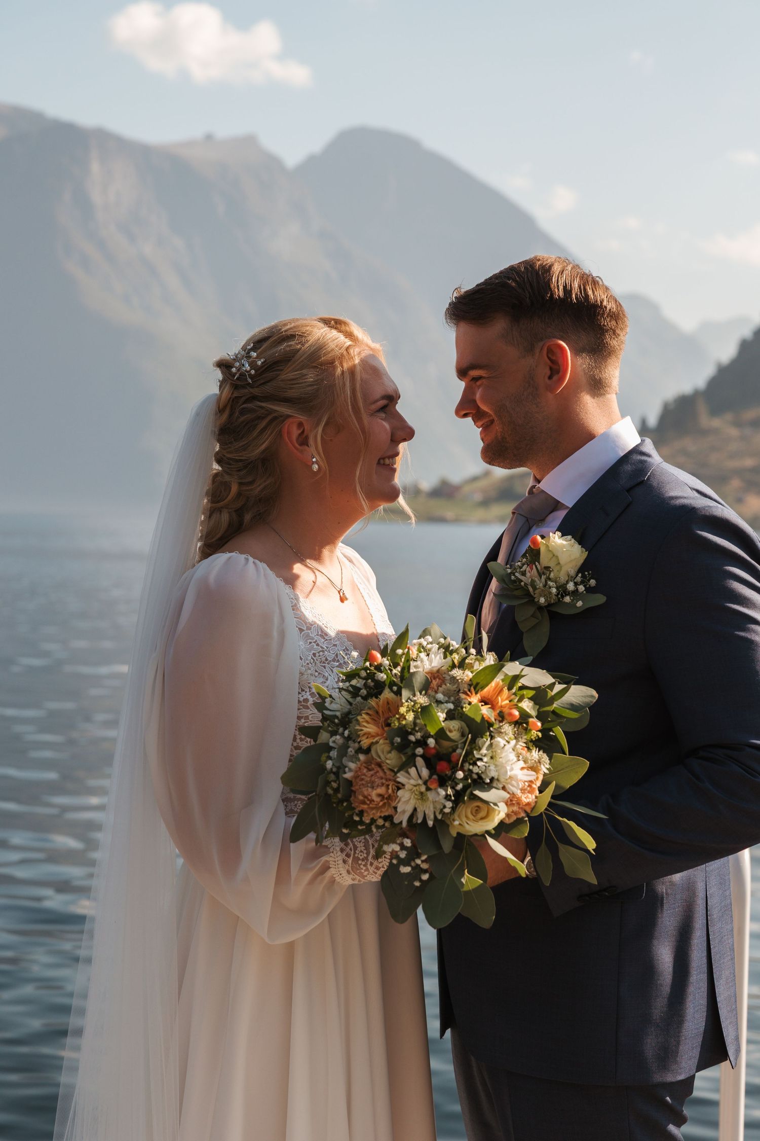 A wedding couple shares an intimate moment by a mountain lake while holding a beautiful peach and white floral bouquet.