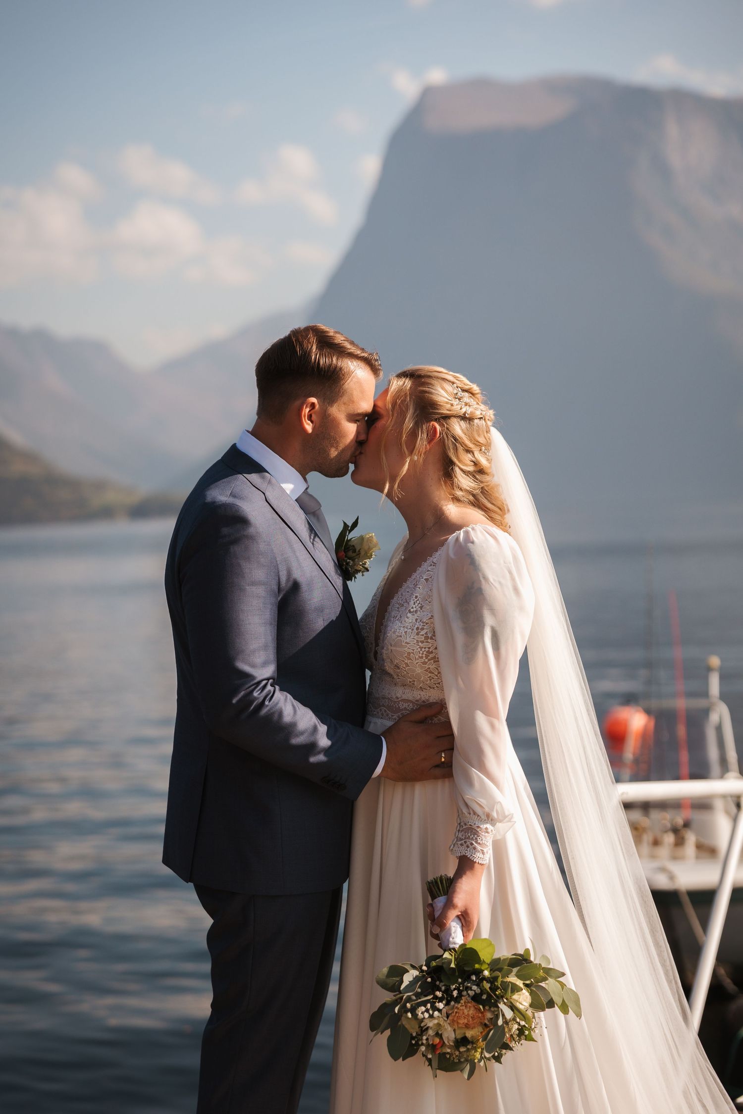 Newlywed couple shares intimate moment by mountain lake with bridal bouquet and scenic backdrop.