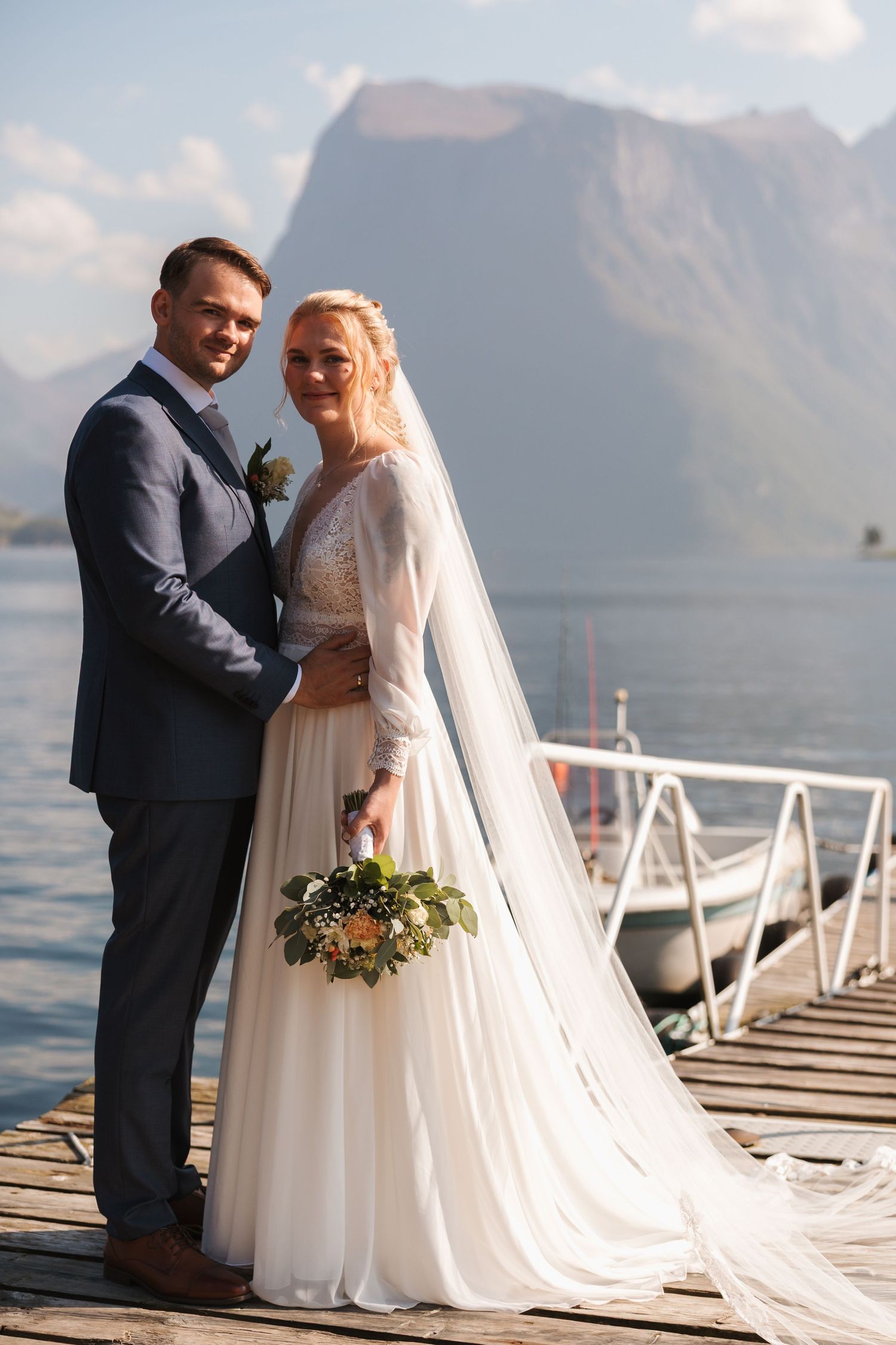 A romantic waterfront wedding couple posing on a dock with mountains and a lake in the background.