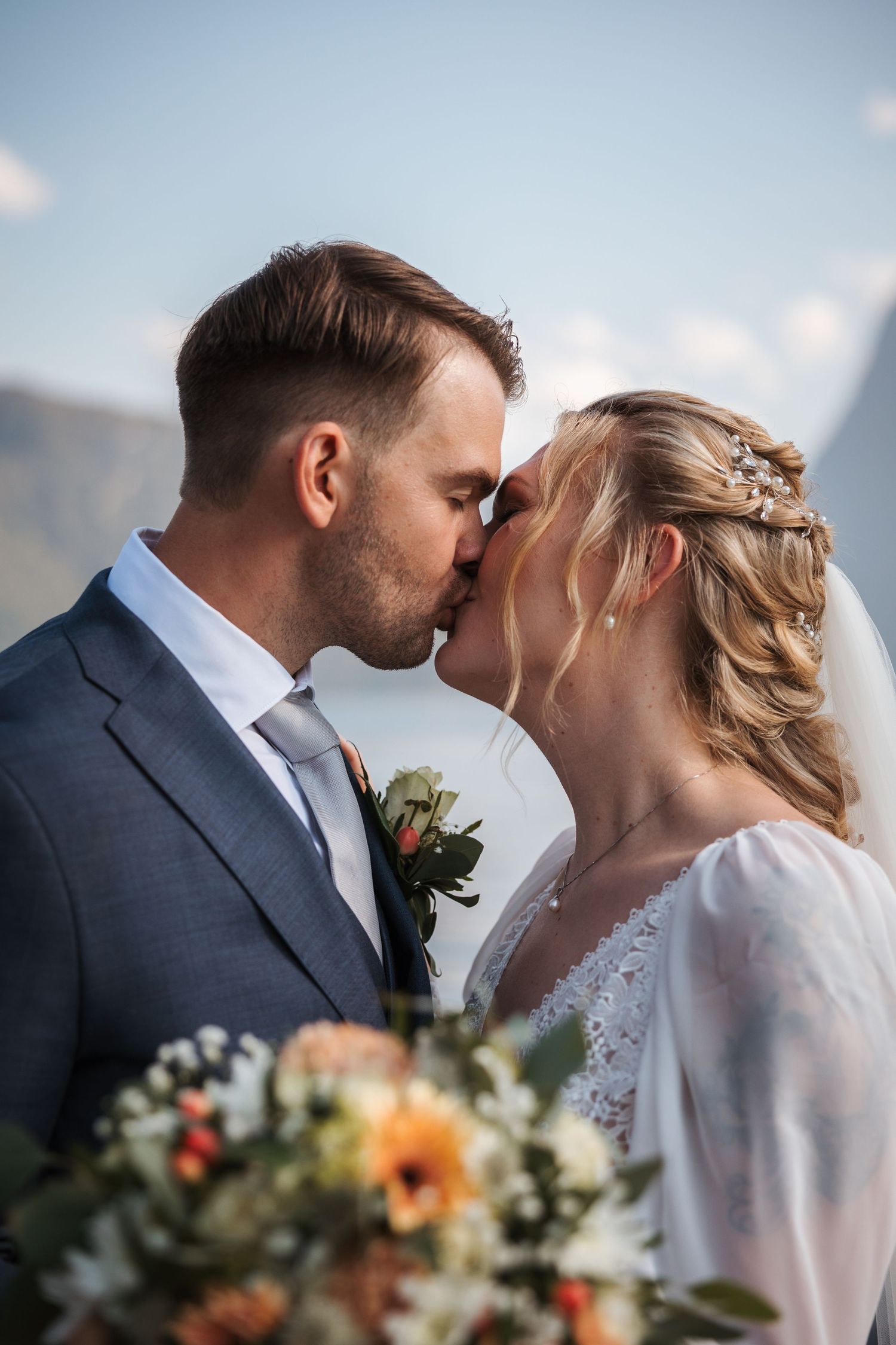 A romantic wedding moment with a couple kissing outdoors, featuring a beautiful floral bouquet with orange and white flowers.