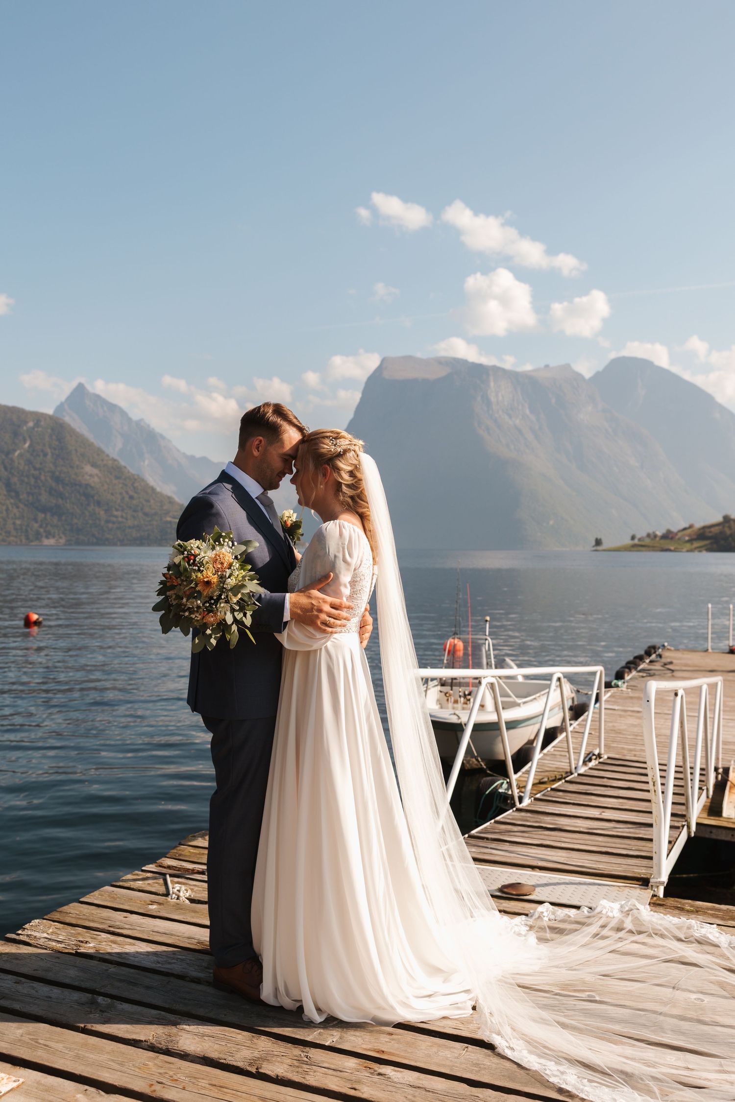 Newlywed couple shares romantic moments on a wooden dock with stunning mountain lake views at a waterfront wedding.