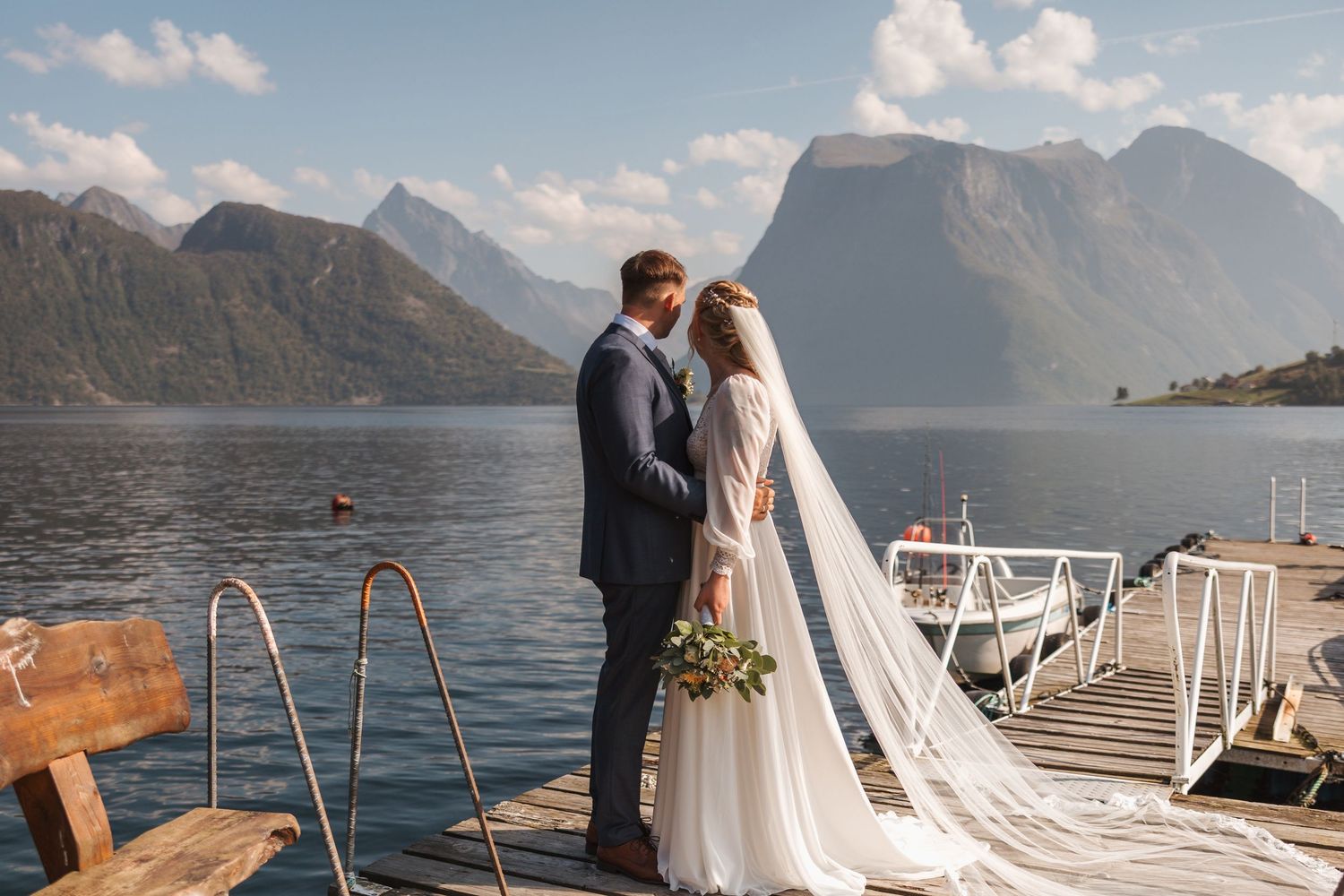 A romantic newlywed couple stands together on a wooden dock overlooking a serene mountain lake and fjord landscape.