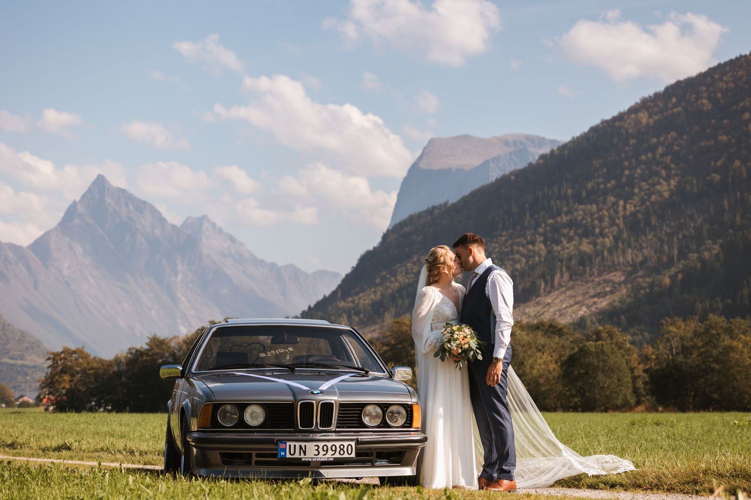 Scenic mountain landscape with wedding couple embracing in foreground and luxury car in majestic alpine meadow setting.