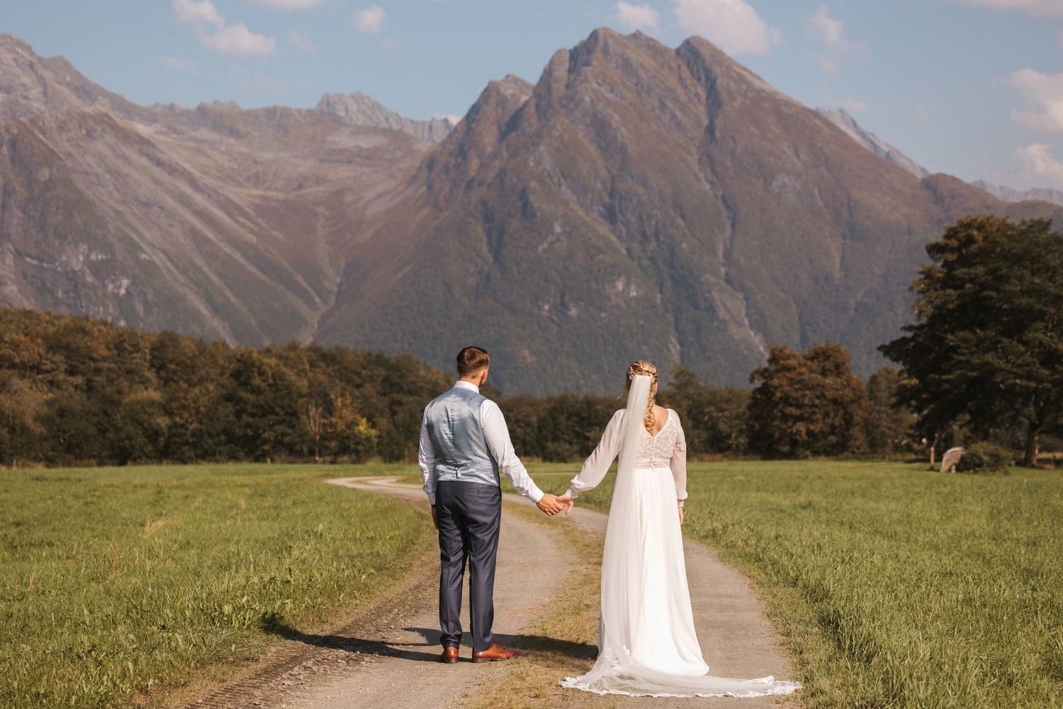 Two people in wedding attire stand along a path with majestic mountain peaks rising dramatically in the background.