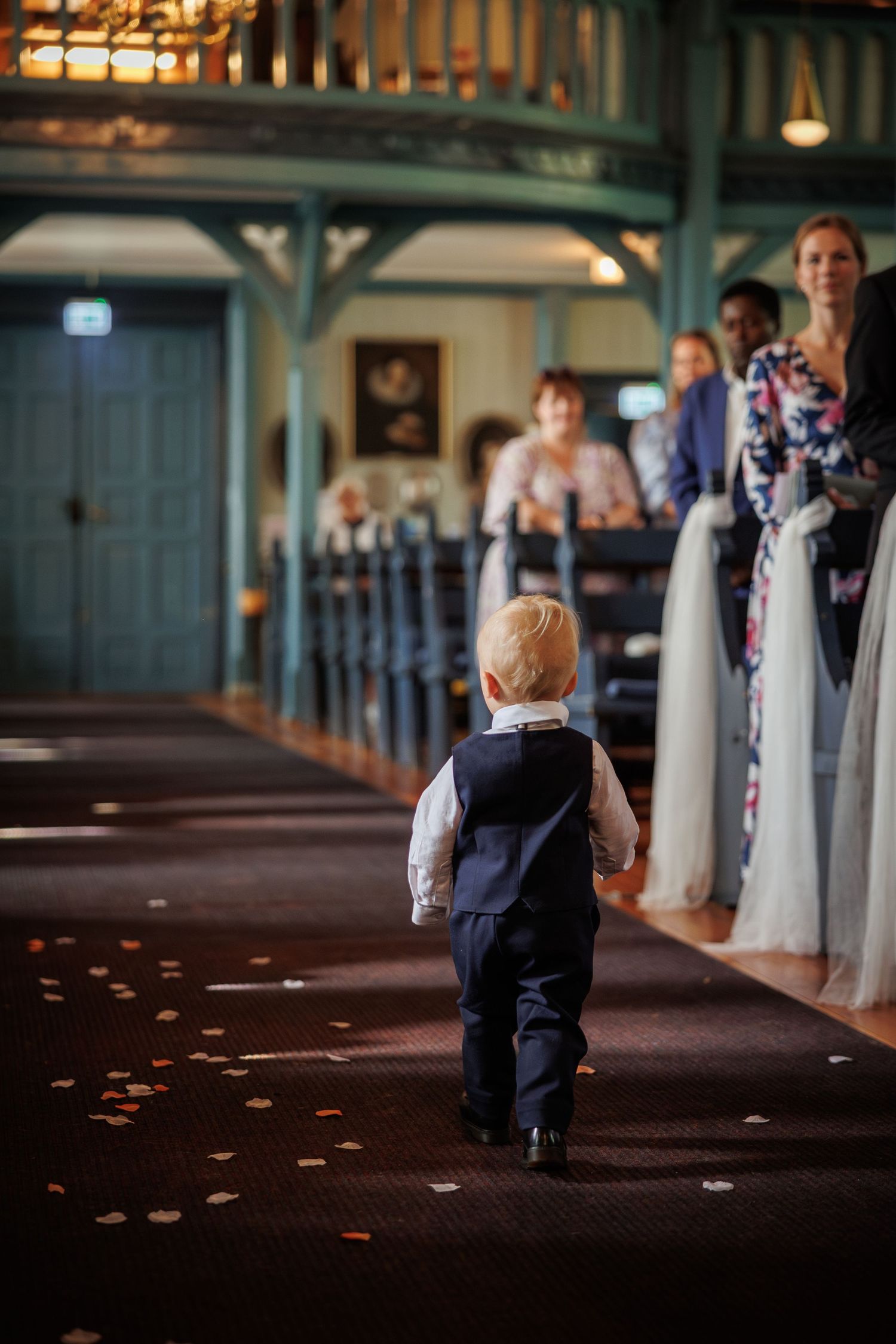 Small boy in formal attire walks down church aisle during wedding ceremony as onlookers watch from pews.