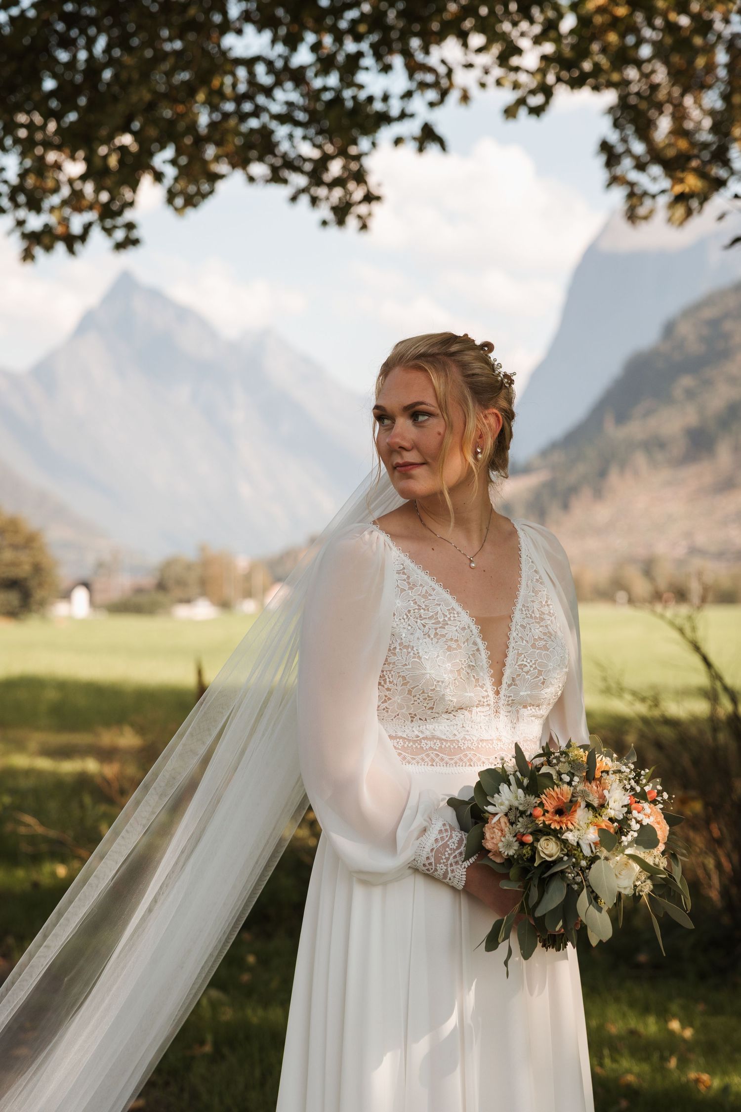 A bride in a flowy white long-sleeved wedding dress holds a bouquet against a backdrop of mountain peaks at sunset.