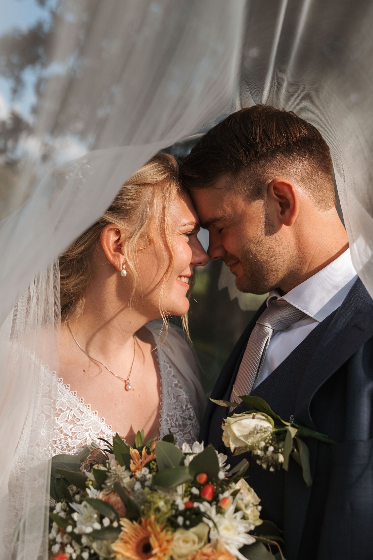 Romantic wedding couple shares an intimate moment with their foreheads touching while holding a rustic floral bouquet.