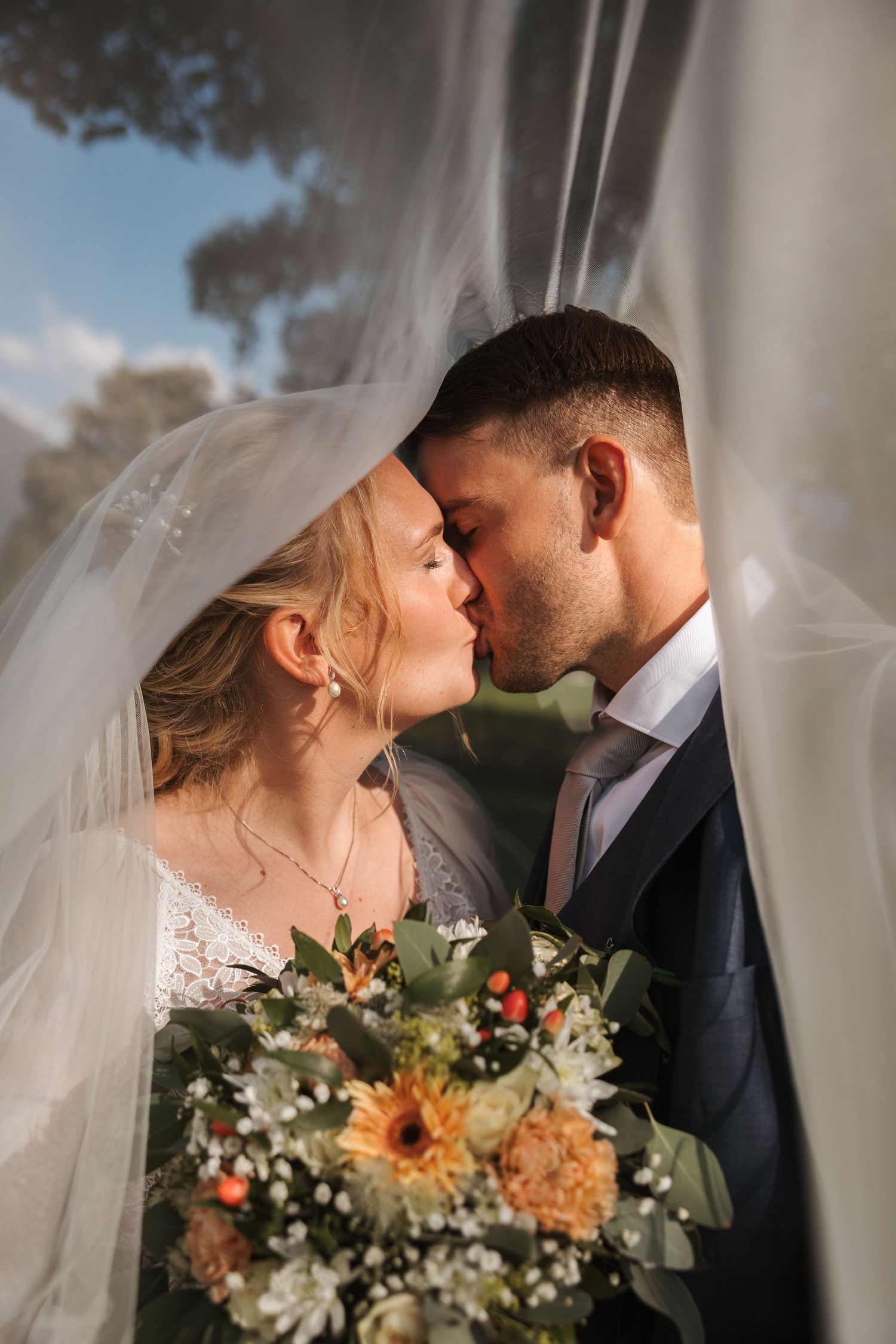 Romantic wedding portrait showing a tender kiss through a flowing veil with autumn bouquet of sunflowers and greenery.